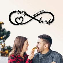 Personalized infinity heart metal sign with custom names and date, laser-cut from black powder-coated steel, displayed against a white brick wall.