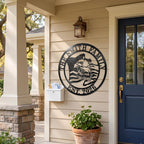 A custom matte black 14-gauge steel eagle flag family monogram is mounted on the beige-sided wall of a traditional American Craftsman front porch, next to a navy blue door. The sign, featuring 'THE SMITH FAMILY' and 'EST. 2026', is positioned as personalized home decor, with potted geraniums and a white mailbox below. Natural sunlight illuminates the scene.