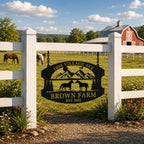 Custom black metal farm sign hanging on a white wooden fence with horses grazing and a red barn in the background.