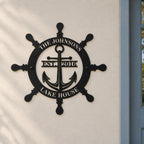 Personalized ship wheel and anchor metal sign mounted on a light wall beside a blue-gray lake house entrance door.