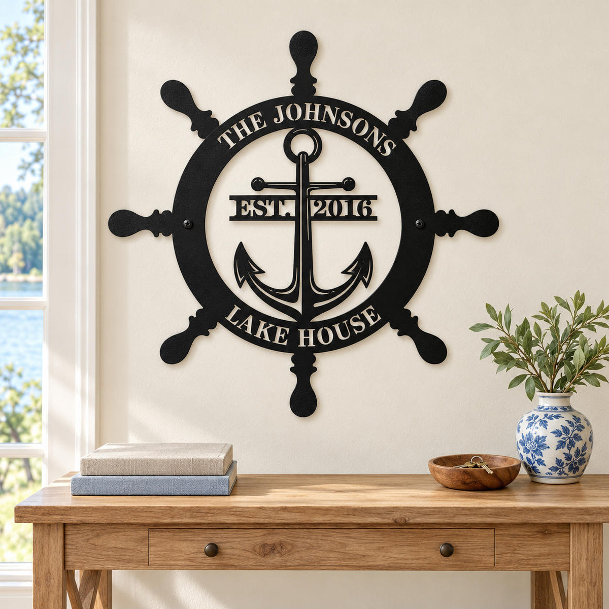 Nautical family metal sign above a wooden console table with books and a blue-and-white vase in a bright lake house entryway.