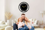 Large personalized black metal wall clock with a solid center dial, featuring laser-cut cutout names David and Linda, established 2023, and Roman numerals showing white wall background.