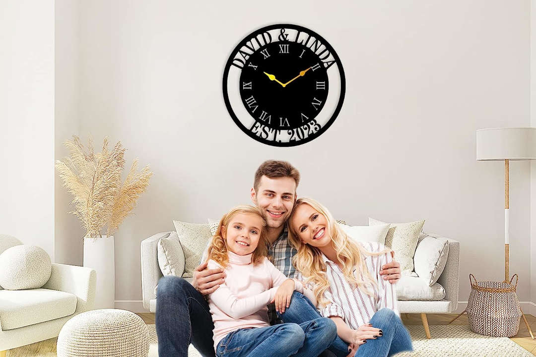 Large personalized black metal wall clock with a solid center dial, featuring laser-cut cutout names David and Linda, established 2023, and Roman numerals showing white wall background.
