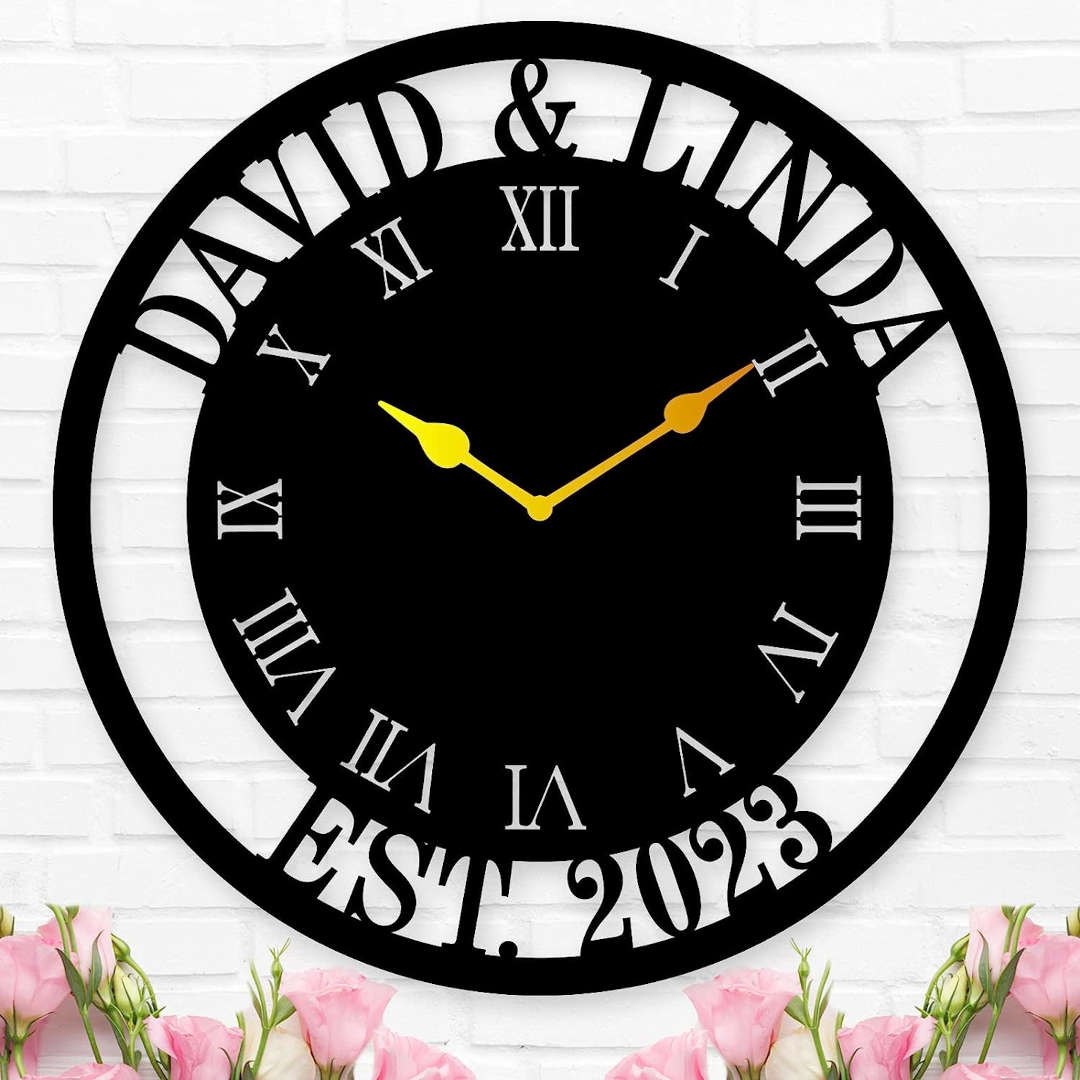 Large personalized black metal wall clock with a solid center dial, featuring laser-cut cutout names David and Linda, established 2023, and Roman numerals showing white wall background. Main image