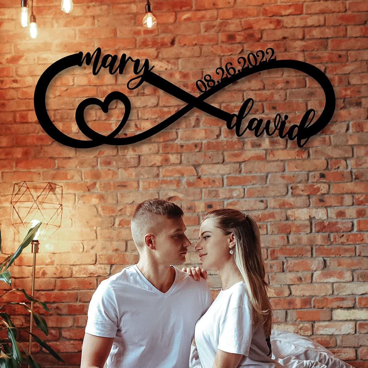 Personalized infinity heart metal sign with custom names and date, laser-cut from black powder-coated steel, displayed against a white brick wall.