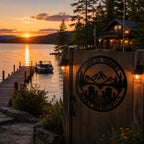 Black metal lake house wall sign mounted near a lakeside dock at sunset with warm golden light and mountain views.