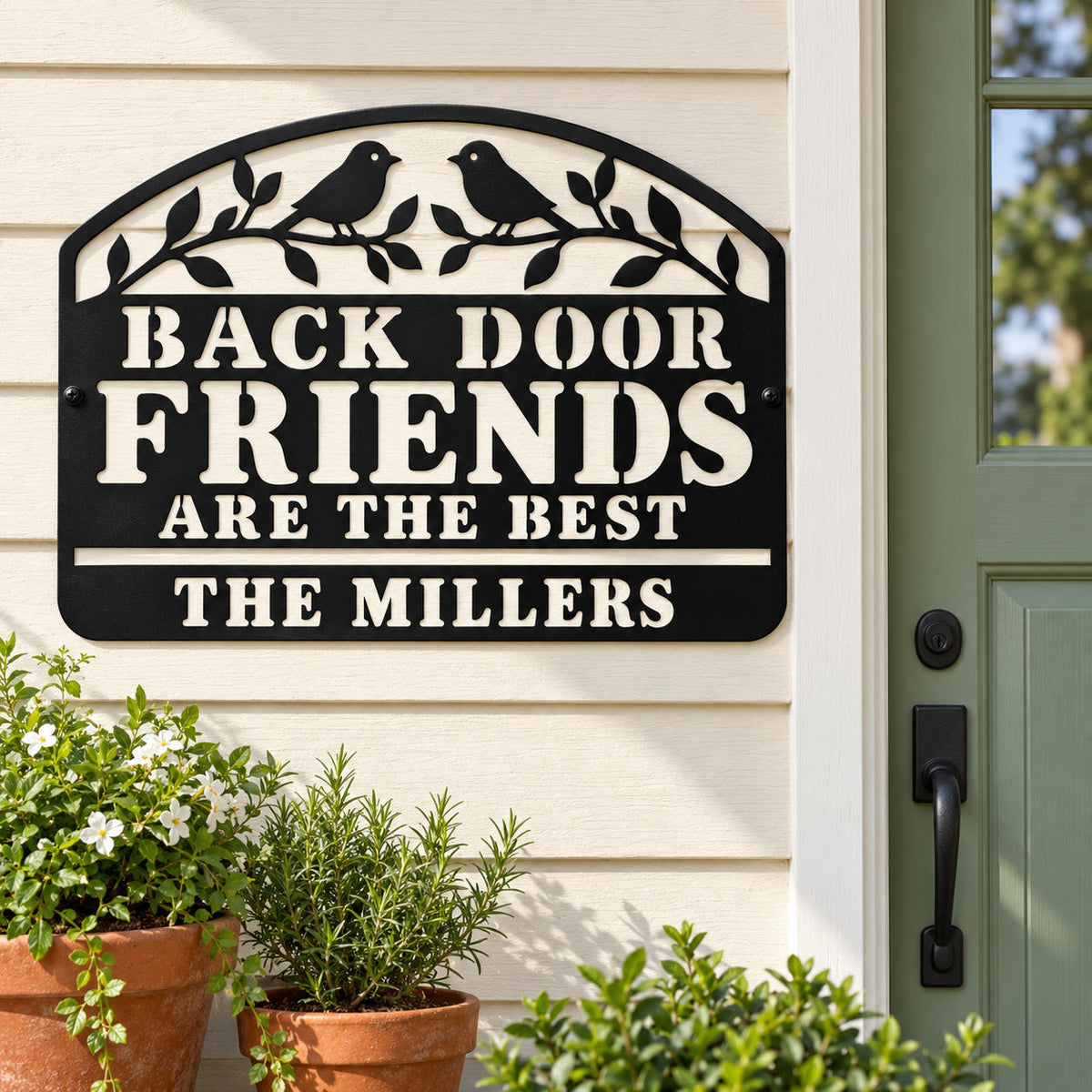 Personalized black metal welcome sign mounted beside a sage green back door with potted plants below.