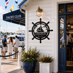 Personalized nautical metal sign displayed at the entrance of a yacht club with marina and sailboats in the background.