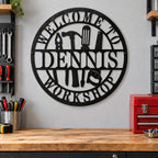 Personalized Dennis workshop metal sign on a gray garage wall above a wooden workbench beside tools and a red toolbox.